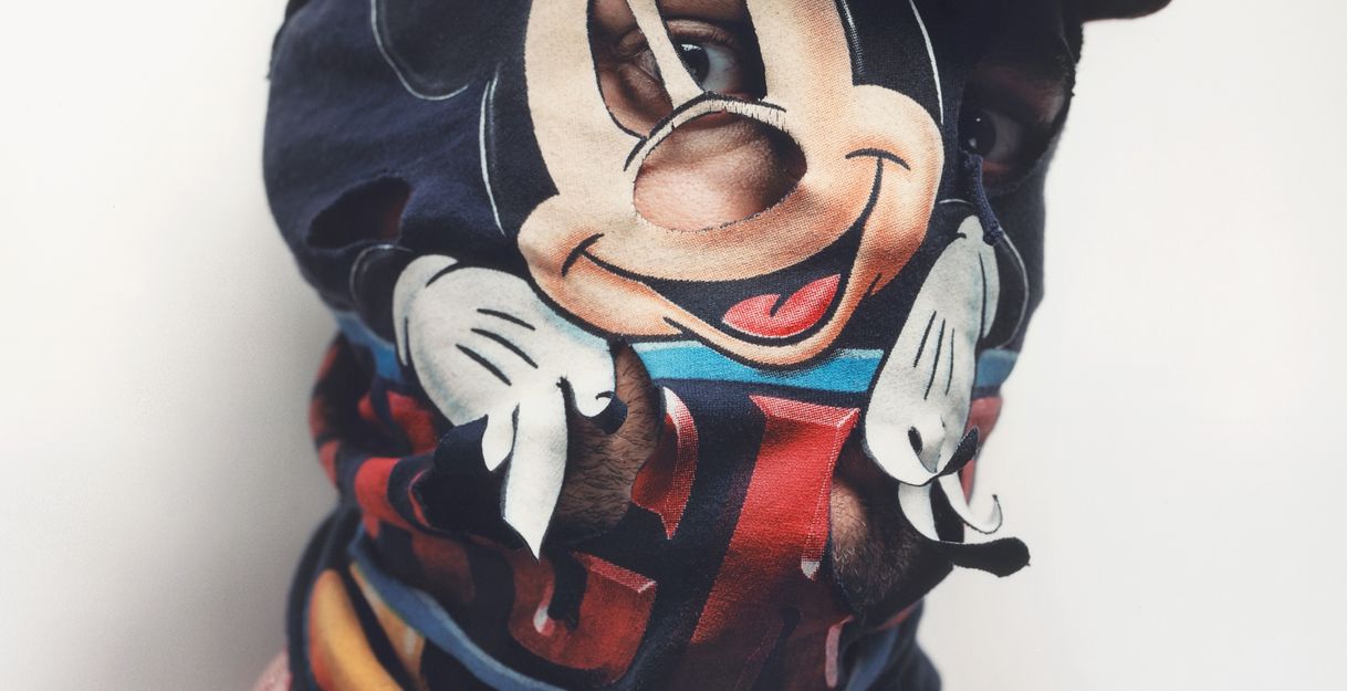 Eine Porträtaufnahme bis zu den Schultern. Das Gesicht ist durch eine weinrote Mütze und vor allem durch eine Stoffmaskierung verdeckt, die die Trickfilmfigur Mickey Mouse zeigt.