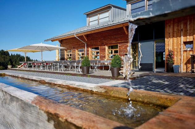 Ein modernes, holzverkleidetes Gebäude mit einer Terrasse und Sonnenschirmen. Im Vordergrund fließt Wasser aus einem Brunnen.