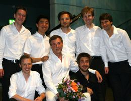 Ein Gruppenbild der jungen Pianisten in weißen Hemden gekleidet. Der Herr in der Mitte sitzend hält einen bunten Blumenstrauß.