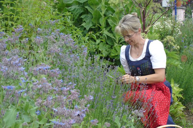 Frau mit Brille und Tracht kniet in einem Kräutergarten, umgeben von blühendem Borretsch und anderen grünen Pflanzen.