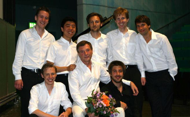 Ein Gruppenbild der jungen Pianisten in weißen Hemden gekleidet. Der Herr in der Mitte sitzend hält einen bunten Blumenstrauß.