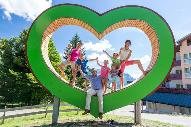 Eine fröhliche Familie posiert in einem großen grünen Herz-Symbol. Im Hintergrund sind Bäume und ein Gebäude zu sehen, bei sonnigem Wetter.