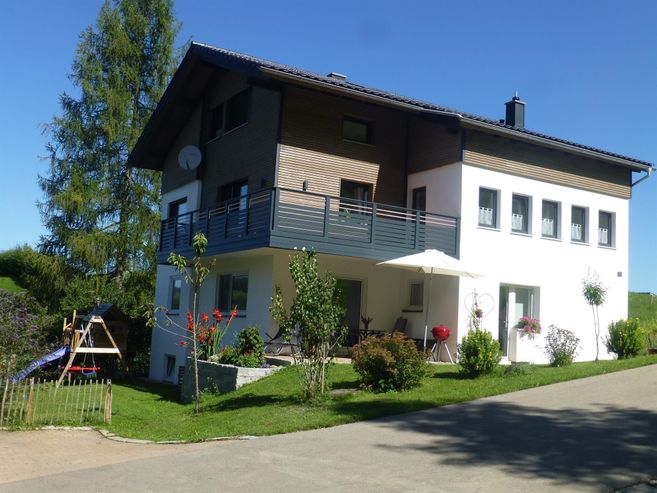 A modern house with two floors, surrounded by a well-maintained garden. In the foreground, there is a terrace with a sun umbrella and flower beds.