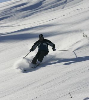 Skifahrer in dunkler Kleidung fährt einen sonnigen, verschneiten Hang hinab.