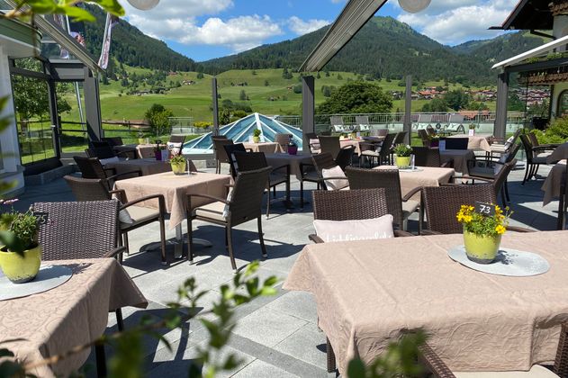 Eine einladende Terrasse mit Tischen und Stühlen, umgeben von einer schönen Landschaft. Im Hintergrund sind Berge und ein klarer Himmel zu sehen.