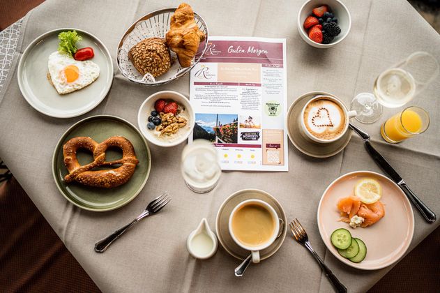 Ein reichhaltiges Frühstück mit verschiedenen Gerichten auf einem Tisch. Zu sehen sind ein Spiegelei, ein Croissant, Brezeln, Obst und Getränke.