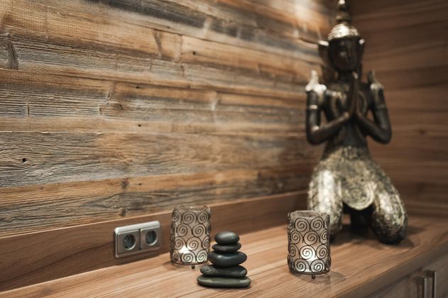 Ein elegantes Dekor mit einem Buddha-Statuette und schwarzen Steinen auf einer Holzoberfläche. Zwei dekorative Kerzenhalter ergänzen das beruhigende Ambiente im Spabereich.