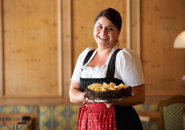 Eine lächelnde Frau trägt eine Torte in traditioneller bayerischer Tracht. Der Hintergrund zeigt eine gemütliche Holzwand und eine einladende Atmosphäre.