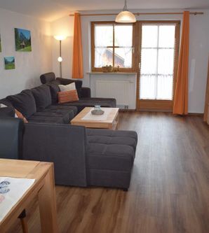 Ein modernes Wohnzimmer mit einer großen Couch und einem Holztisch. Helle Fenster mit Vorhängen und ein Fernseher auf der gegenüberliegenden Wand.