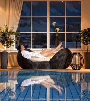 Eine entspannte Frau in einem weißen Bademantel liegt auf einem modernen Liegesessel am Rand eines Schwimmbeckens. Große Fenster bieten einen Blick auf die winterliche Landschaft.
