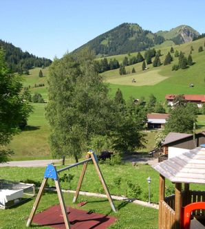 Eine Spielplatzlandschaft mit einer Schaukel und einem kleinen Spielhaus. Im Hintergrund sind grüne Hügel und Berge zu sehen, die von Bäumen umgeben sind.