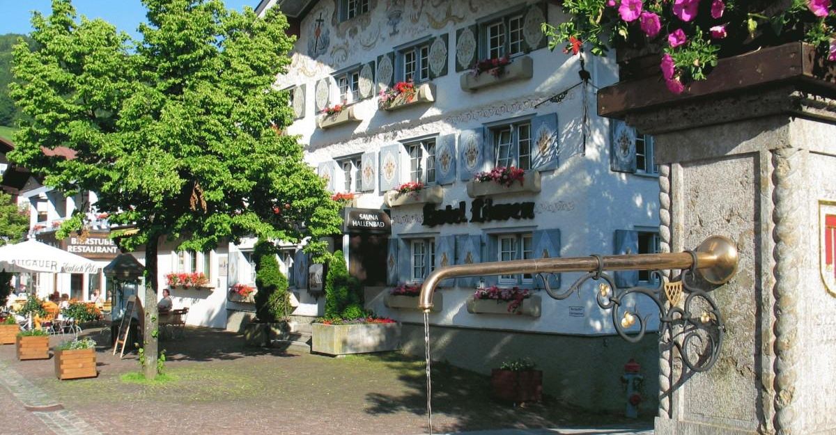 Ein charmantes Gebäude mit bunten Fensterläden und Blumenkästen. Im Vordergrund befindet sich ein Brunnen und grüne Bäume.