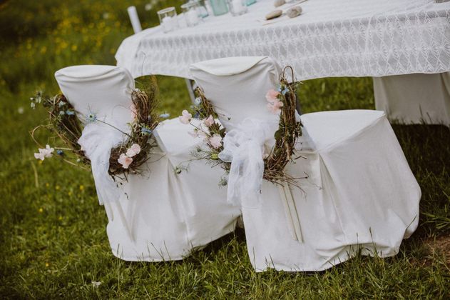 Zwei elegante, mit Blumen und Dekoration geschmückte Stühle stehen an einem festlich gedeckten Tisch. Die Umgebung ist grün und natürlich, ideal für eine Feier im Freien.