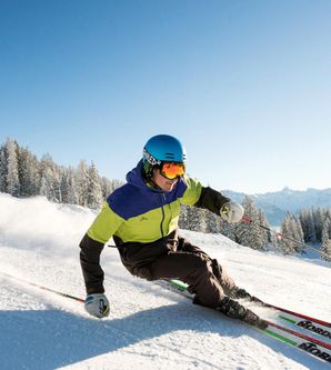 Skifahrer in bunten Skianzug fährt verschneite Piste hinab, stützt sich mit Skistöcken ab.