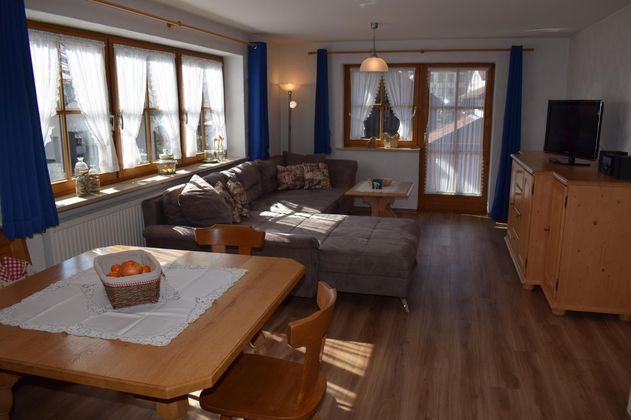 Ein gemütlicher Wohnraum mit einer großen Couch und einem Esstisch. Helle Fenster und freundliche Einrichtung schaffen eine einladende Atmosphäre.