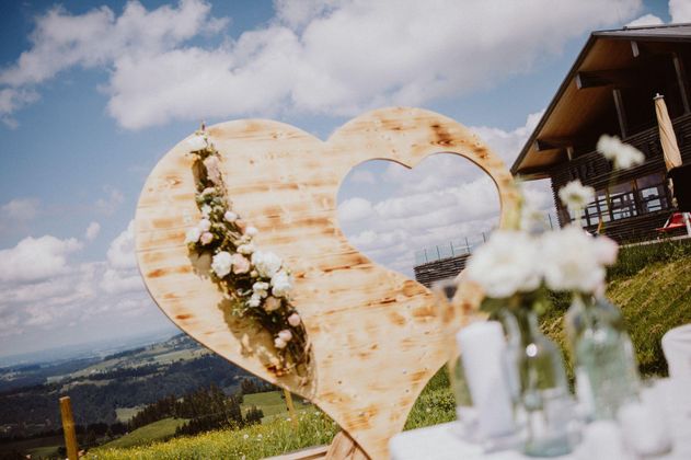 Ein dekoriertes Holzherz mit Blumen, das vor einer malerischen Landschaft steht. Im Hintergrund sind Wolken und ein Gebäude sichtbar.