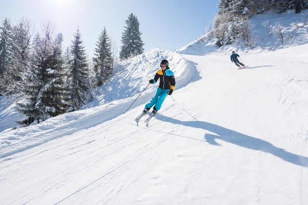 Zwei Skifahrer auf sonniger, verschneiter Piste vor bewaldetem Hang.