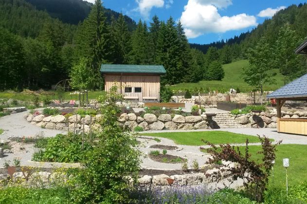Weitläufiger Kräutergarten mit verschiedenen Beeten, Steinmauern, Wegen, Holzhäusern und grünen Bäumen vor bergiger Landschaft.