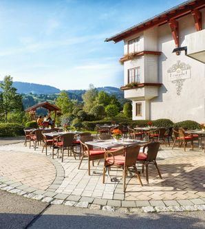 Eine einladende Terrasse mit Tischen und Stühlen umgeben von schöner Landschaft. Im Hintergrund ist ein charmantes Gebäude zu sehen, das eine gemütliche Atmosphäre schafft.
