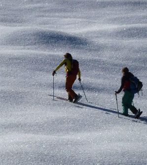 Zwei Personen beim Skilanglauf in einer weiten, hügeligen Schneelandschaft. Lange Schatten der Skiläufer auf dem glitzernden Schnee.