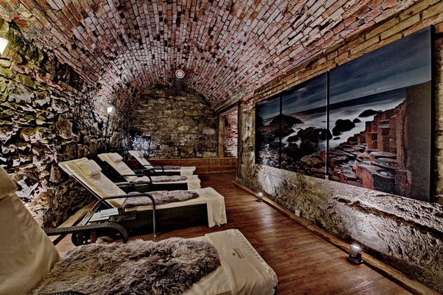 Ein entspannender Wellnessraum mit gemütlichen Liegen und einer steinernen Wand. An der Wand hängt ein großes Bild einer Landschaft.