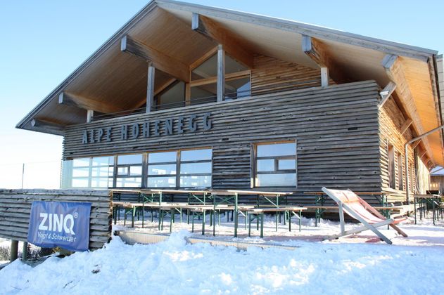 Eine gemütliche Berghütte aus Holz mit großen Fenstern. Vor der Hütte liegt Schnee und es stehen einige Tische und Stühle im Freien.