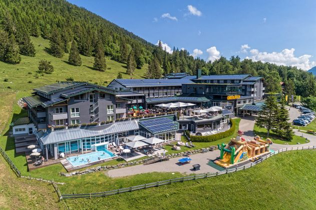Ein modernes Hotel in den Bergen mit einem Pool und einem Spielplatz. Die Umgebung ist grün und von Bäumen umgeben.