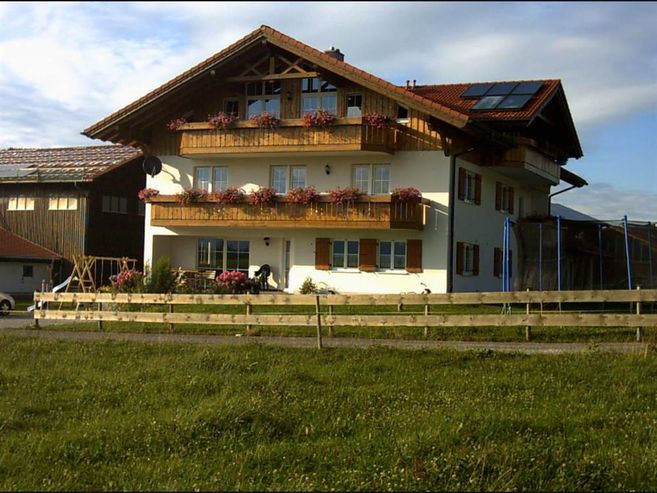 A traditional Bavarian house with several balconies and flowers. The building is situated in a rural setting with meadows and a hangar in the background.