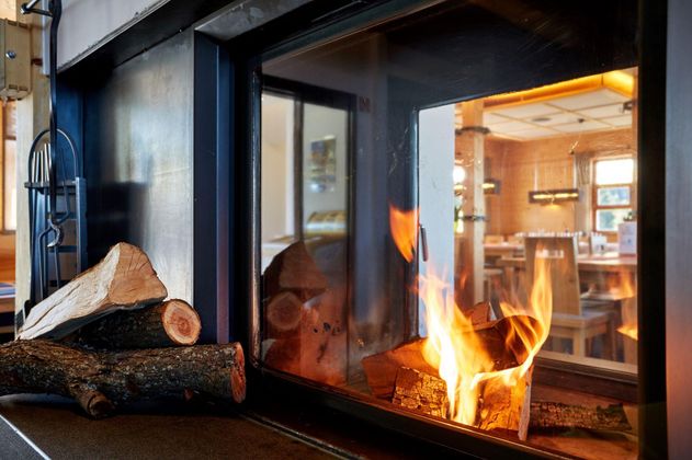 Ein gemütlicher Kamin mit loderndem Feuer und Holzscheiten. Im Hintergrund ist ein einladender Raum zu sehen.