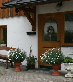 Ein gemütlicher Eingang mit einer Holzbank und blühenden Pflanzen in Töpfen. Die Fassade ist schlicht und einladend.