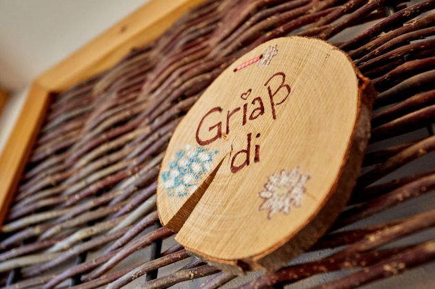 Ein Holzschild mit der Aufschrift „Griaß di“ und dekorativen Blüten. Es hängt an einer geflochtenen Wand.