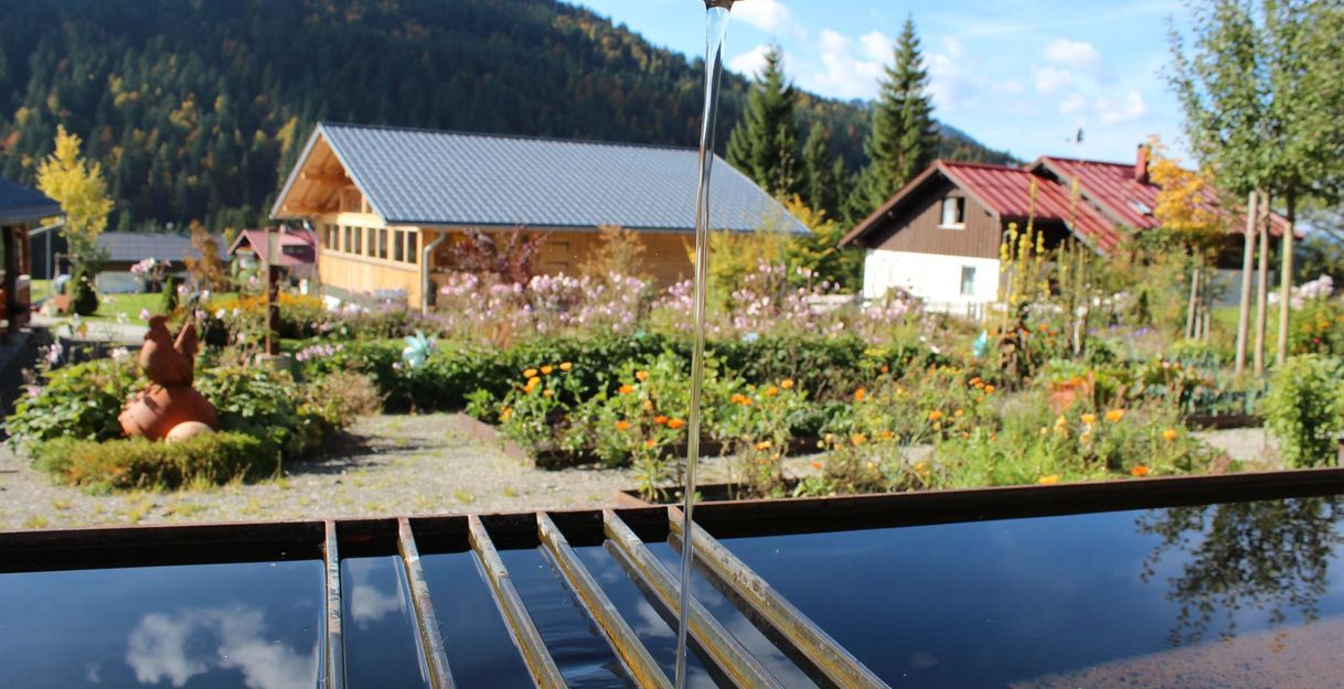 Wasserstrahl aus Metallrohr fällt in dunkles Wasserbecken, bunte Blumenbeete mit Häusern im Hintergrund.