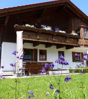 Ein schönes Holzhäuschen mit einem Balkon und blühenden Blumen im Vordergrund. Die Umgebung ist grün und die Sonne scheint.