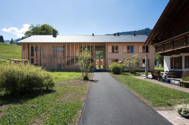 Ein modernes Haus mit Holzverkleidung und Glasfront in einer ruhigen Umgebung. Der Weg führt durch einen gepflegten Garten mit grünen Pflanzen.