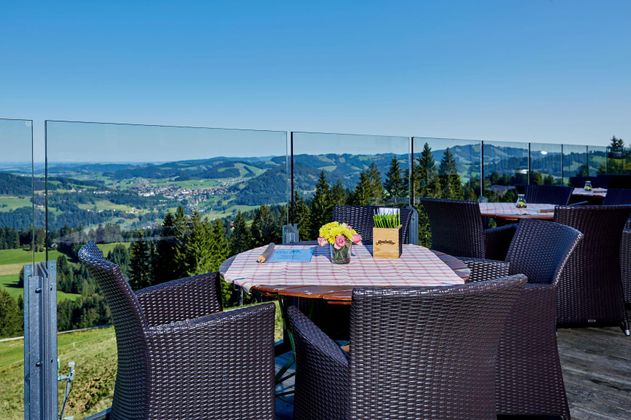 Ein gemütlicher Tisch mit Stühlen auf einer Terrasse mit Blick auf eine wunderschöne Berglandschaft. Die klare, blaue Himmel und das grüne Tal schaffen eine einladende Atmosphäre.