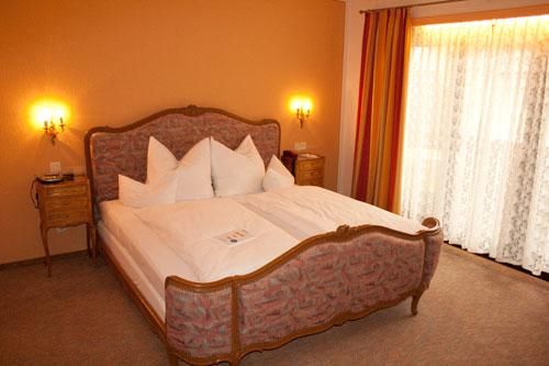 Ein gemütliches Schlafzimmer mit einem großen Bett und dekorativen Kissen. Weiche Beleuchtung und Vorhänge sorgen für eine einladende Atmosphäre.