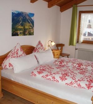 Ein gemütliches Schlafzimmer mit einem Doppelbett und rot gemusterten Kissen. Es gibt Holzmöbel und einen Blick auf die Berge durch das Fenster.