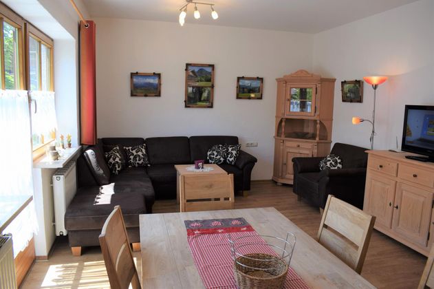 Ein gemütliches Wohnzimmer mit einem schwarzen Sofa und einem Holztisch. An den Wänden hängen Bilder, und es gibt einen Fernseher sowie eine elegante Vitrine.