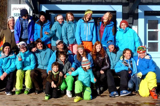 Große Gruppe von Personen in Winterkleidung posiert vor einem Gebäude mit blauen Türen und Fensterläden. Viele tragen blaue Jacken. Ski lehnen an der Seite. Sonniger Tag.
