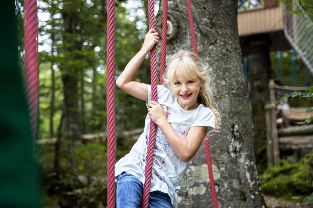 Ein Mädchen schwingt fröhlich an Seilen. Im Hintergrund sind Bäume und ein Spielplatz.