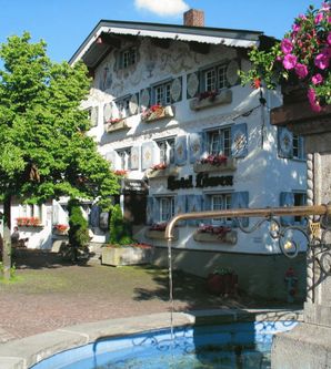 Ein charmantes Gebäude mit bunten Fensterläden und Blumenkästen. Im Vordergrund befindet sich ein Brunnen und grüne Bäume.