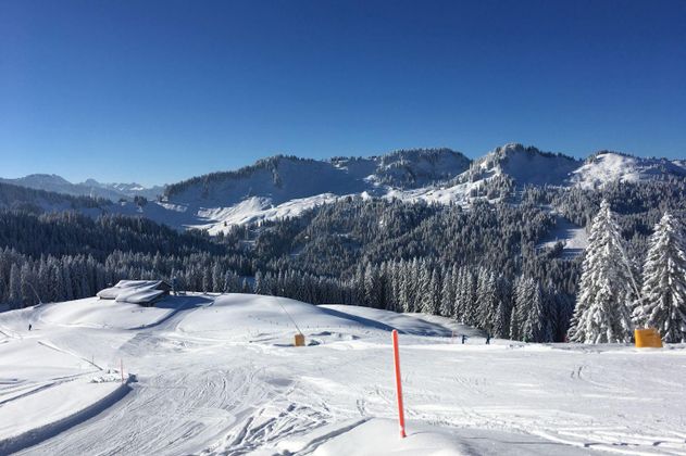 Weite, verschneite Berglandschaft unter strahlend blauem Himmel, einzelne Skifahrer erkennbar.