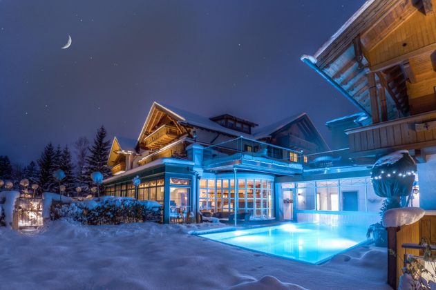 Ein wunderschönes Haus in der Winterlandschaft, beleuchtet von blauen Lichtern. Der Garten ist mit Schnee bedeckt und der Himmel zeigt den Mond in einer klaren Nacht.