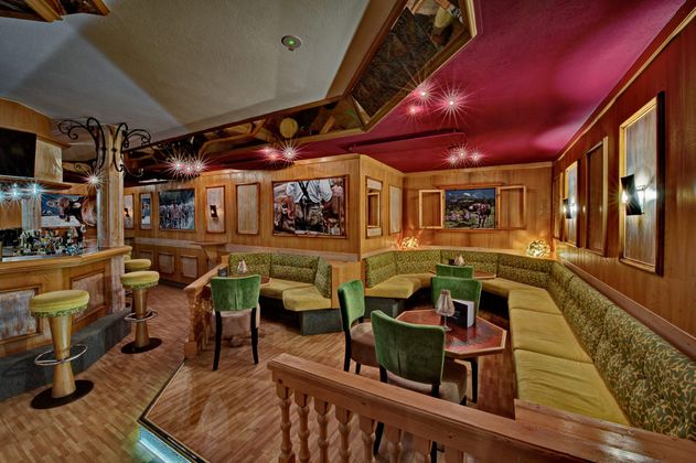 Eine gemütliche Bar mit Holzverkleidung und dekorativen Bildern an den Wänden. Die Sitzbereiche sind mit grünen und gelben Möbeln ausgestattet und die Beleuchtung sorgt für eine einladende Atmosphäre.
