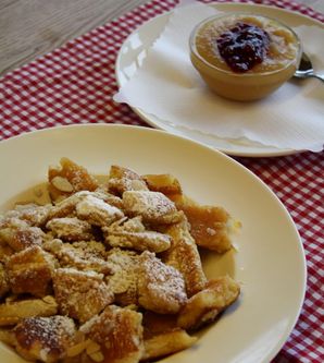Ein Teller mit Kaiserschmarrn, bestäubt mit Puderzucker, liegt auf einem karierten Tischset. Daneben befindet sich ein Schälchen mit Apfelmus und einem Klecks Marmelade.