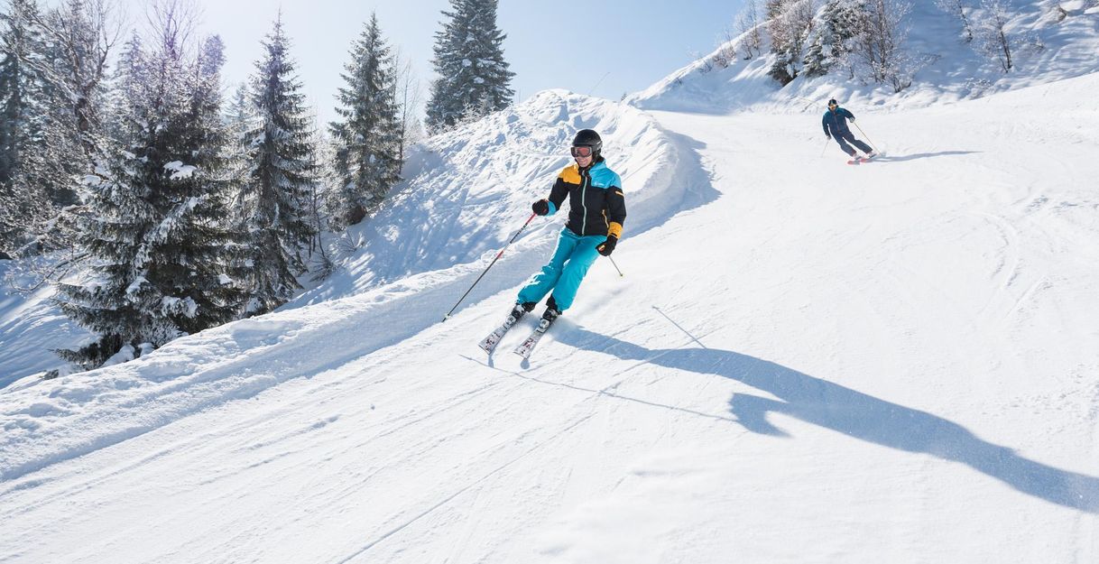 Zwei Skifahrer auf sonniger, verschneiter Piste vor bewaldetem Hang.