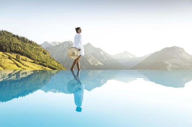 Eine Frau mit einem Hut steht am Rand eines Infinity-Pools. Im Hintergrund sind Berge und ein klarer Himmel sichtbar.