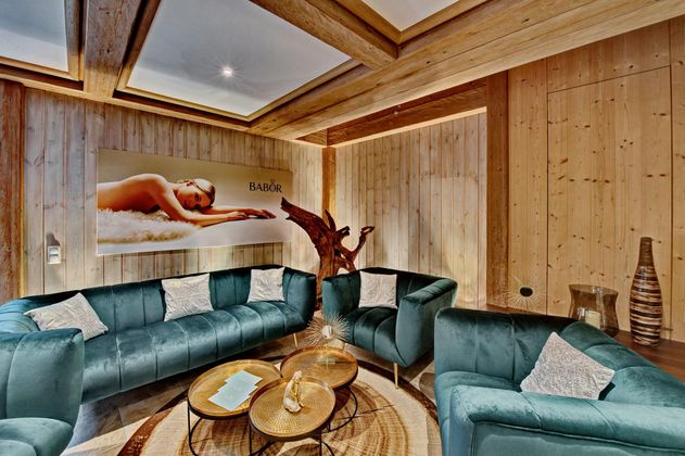 Ein eleganter Lounge-Bereich mit bequemen grünen Sofas und einem stilvollen Holzwand-Design. Im Hintergrund ist ein entspannendes Bild einer Person beim Wellness zu sehen.