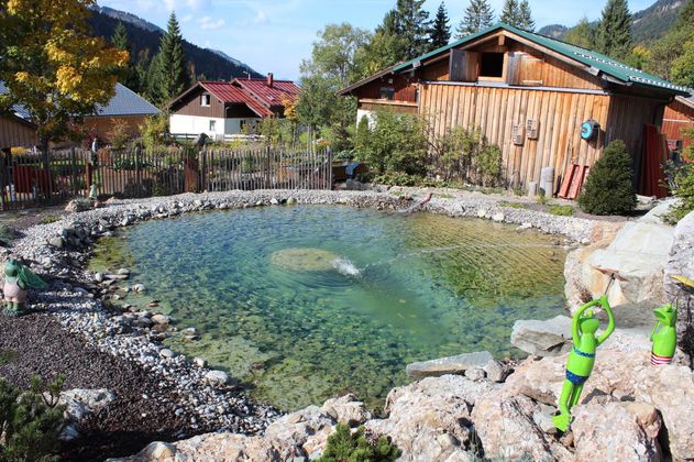 Klarer Teich mit Steinufer, umgeben von bunten Blumen, Holzhäusern und grünen Bäumen.