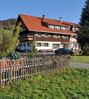 Ein typisches bayerisches Landhaus mit rotem Dach und schöner Umgebung. Im Vordergrund ein Garten mit einem Zaun aus Holz.